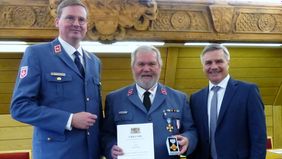 Drei Männer stehen nebeneinander, der Mann in der Mitte in Malteserkleidung hält eine Urkunde und eine Medaille in der Hand.