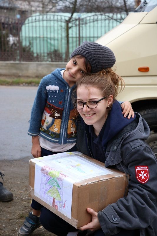Ein Kind mit einer dicken bunten Strickjacke und einer blauen Mütze schmiegt sich lächelnd an eine kniende junge Frau mit einem Paket in der Hand