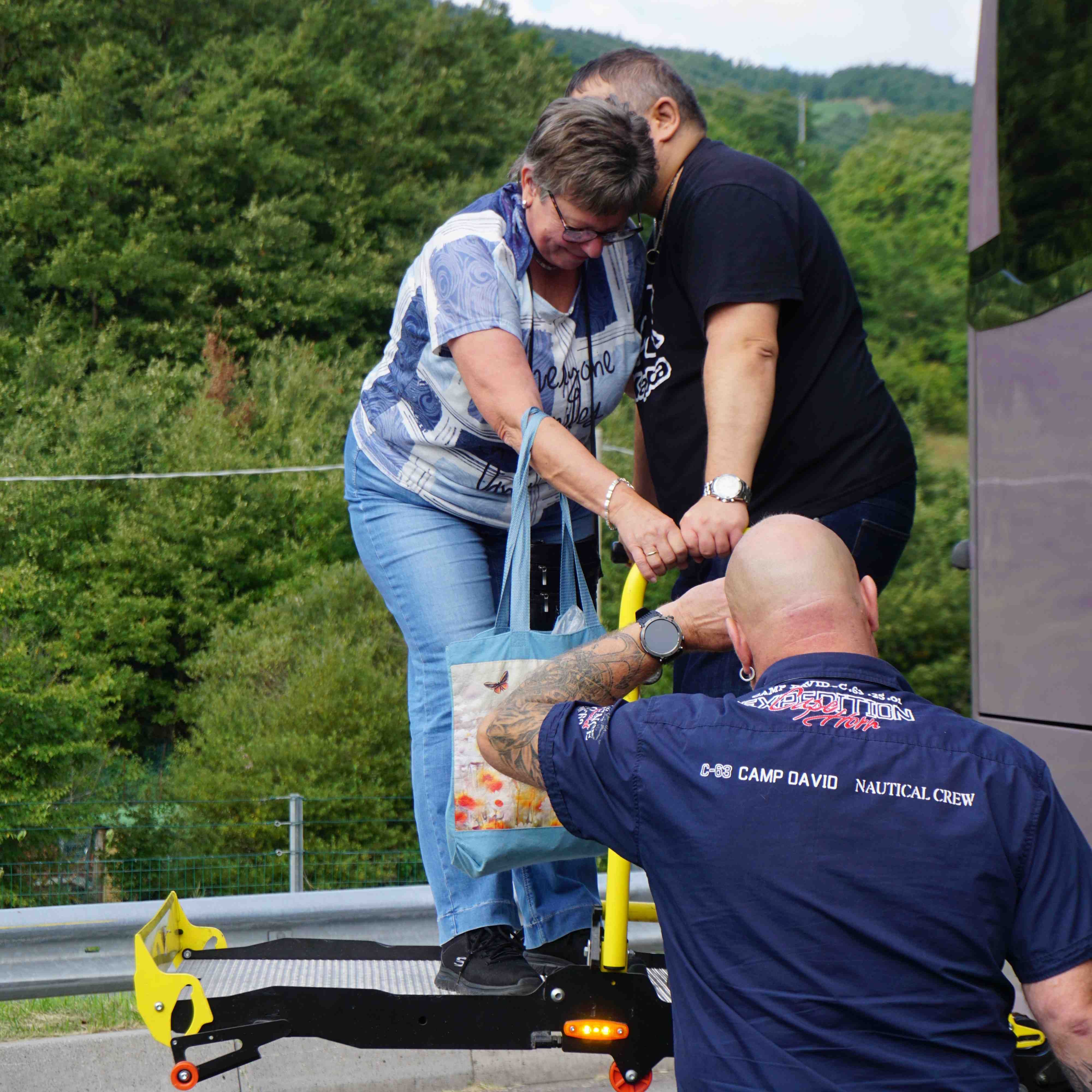 Drei Personen an einer gelben Treppe im Freien, eine trägt eine blaue Jeans und eine Tasche mit Blumenmuster, eine andere eine schwarze Hose und ein schwarzes T-Shirt, die dritte Person trägt ein blaues T-Shirt.
