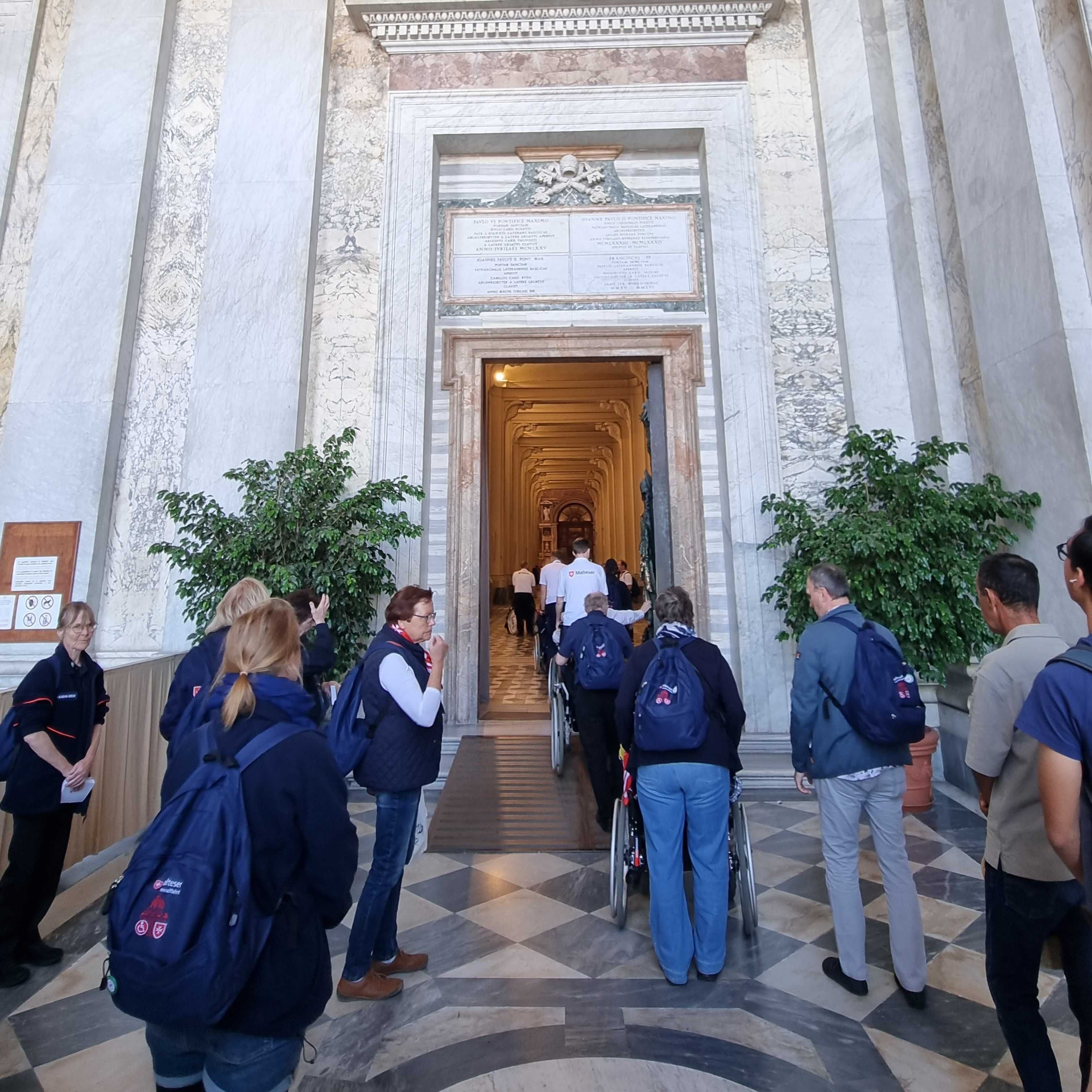 Menschen stehen in einer Reihe vor der Heiligen Pforte im Lateran, umgeben von weißen Steinwänden und zwei großen Pflanzen in Töpfen.