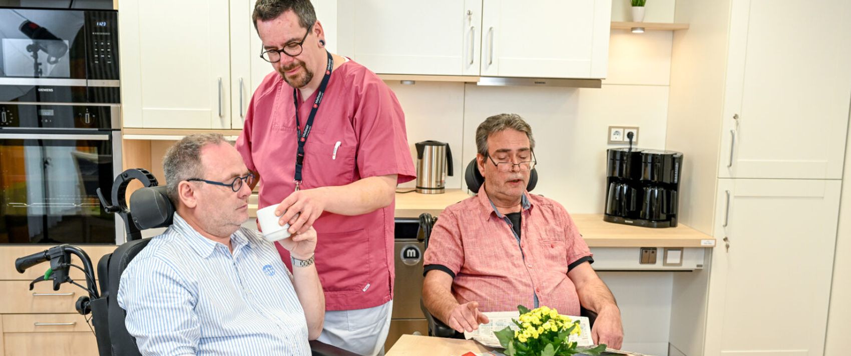 Zwei Patienten im Rollstuhl sitzen am Tisch und ein Pflegerin hilft einem beim Trinken aus einer Tasse
