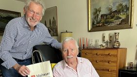 Paul Breitner lehnt hinter einem Sessel, auf dem ein älterer Mann sitzt. Beide lächeln.