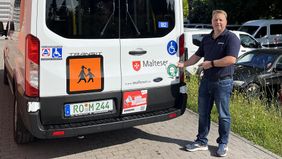 Ein Mann zeigt auf einen Aufkleber an einem Spezialfahrzeug für die Beförderung im Rollstuhl.