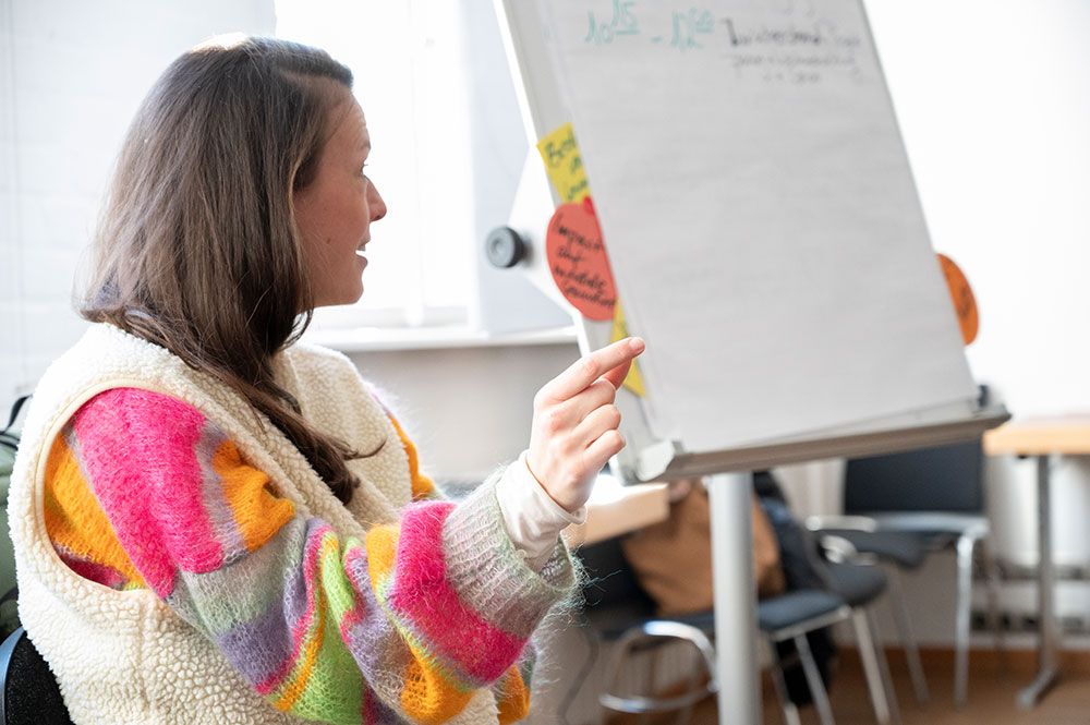 Imke Siefer sitzt auf einem Stuhl und zeigt auf ein Flipchart. Der Raum wirkt wie ein Seminar- oder Besprechungsraum mit Tischen und Stühlen im Hintergrund.