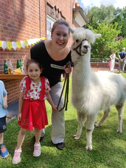 Ein Kind und eine erwachsene Frau stehen gemeinsam mit einem Alpaka im Garten und lächeln