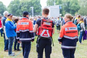 Zu sehen sind zwei Sanitäter sowie eine Sanitäterin von hinten in Einsatzkleidung vor der Kulisse einer Menschenmenge, die den DKT besucht. 