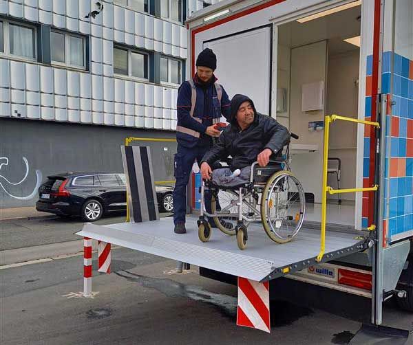 Ein Mann hilft einem Rollstuhlfahrer auf der Laderampe eines Dusch-LKWs