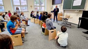 Kinder sitzen in einem Klassenraum. Eine Frau erklärt ihnen etwas.