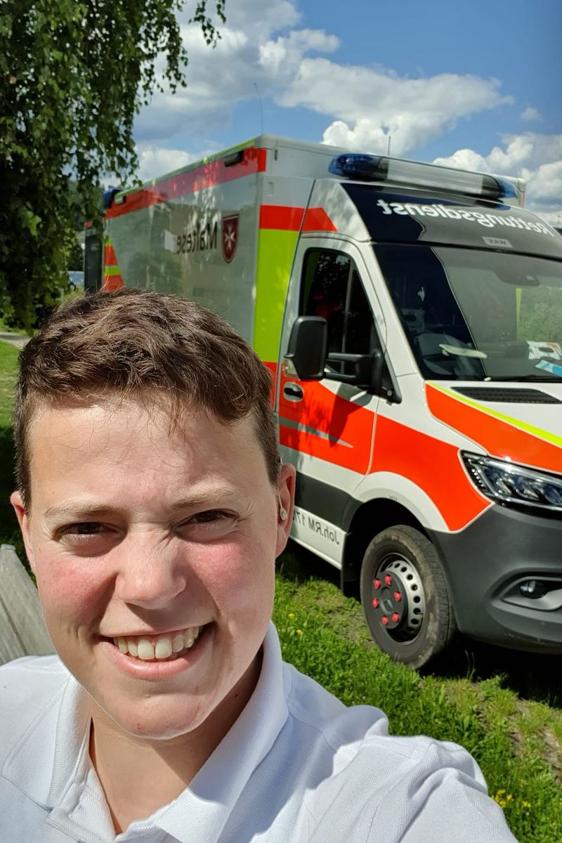 Eine lächelnde junge Frau mit Kurzhaarschnitt vor einem RTW der Malteser.