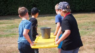 Vier Kinder tragen eine rote Rettungsbare aus Kunststoff. Darauf steht ein Eimer mit Wasser, welchen sie als Geschicklichkeitstest von A nach B tragen müssen.