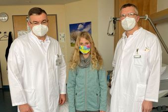 Zwei Ärzte und ein Kind mit Maske stehen in einem Patientenzimmer vor einem Klinikbett.
