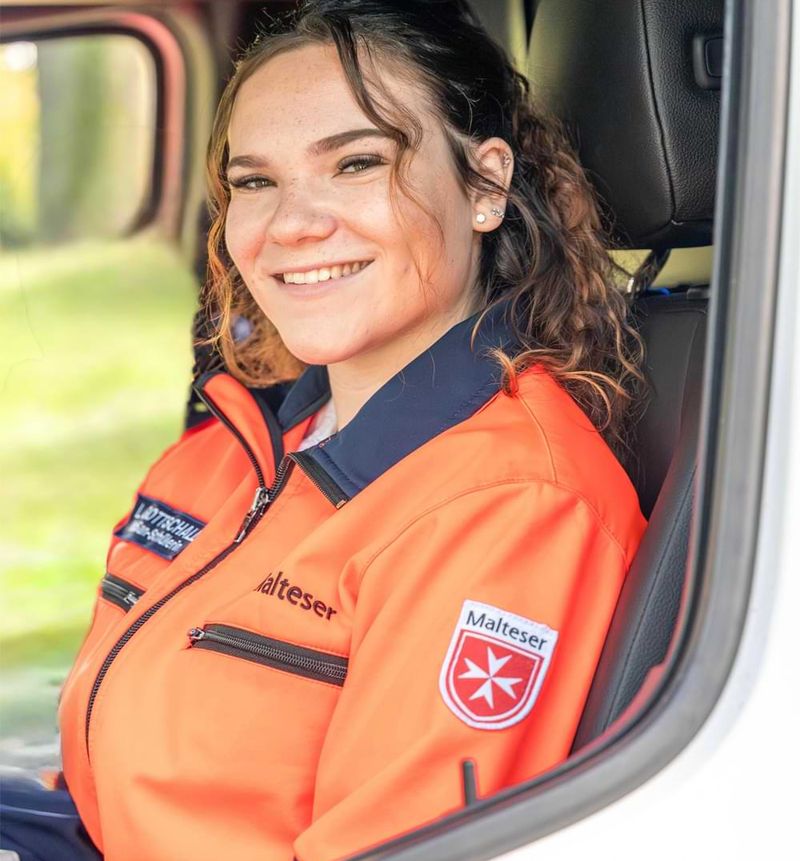 Eine junge Frau in einer leuchtend orangefarbenen Einsatzjacke der Malteser sitzt lächelnd auf dem Fahrersitz eines Einsatzfahrzeugs. Sie blickt freundlich aus dem geöffneten Fenster, während im Hintergrund eine grüne, sonnige Umgebung zu sehen ist.