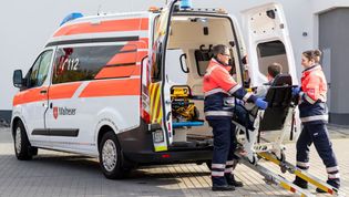 Malteser in Einsatzkleidung helfen einem gehbehindertem Mann in in ein Fahrzeug.