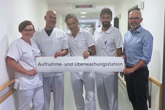 Fünf Klinikmitarbeitende stehen im Flur und halten ein Schild mit der Aufschrift „Aufnahme- und Überwachungsstation“.