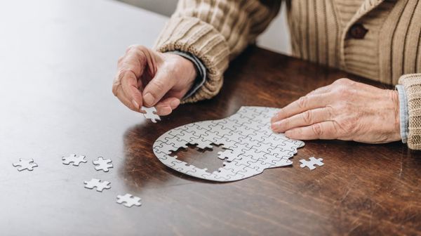 Älterer Mensch sitzt an einem Holztisch und setzt ein kopfförmiges Puzzle zusammen.