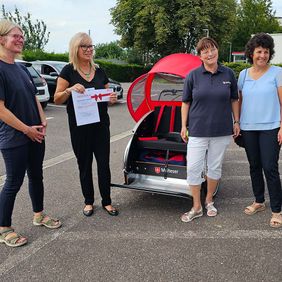 Vier Frauen stehen neben einer Fahrrad-Rikscha. Eine Frau hält ein Zertifikat und einen Gutschein.