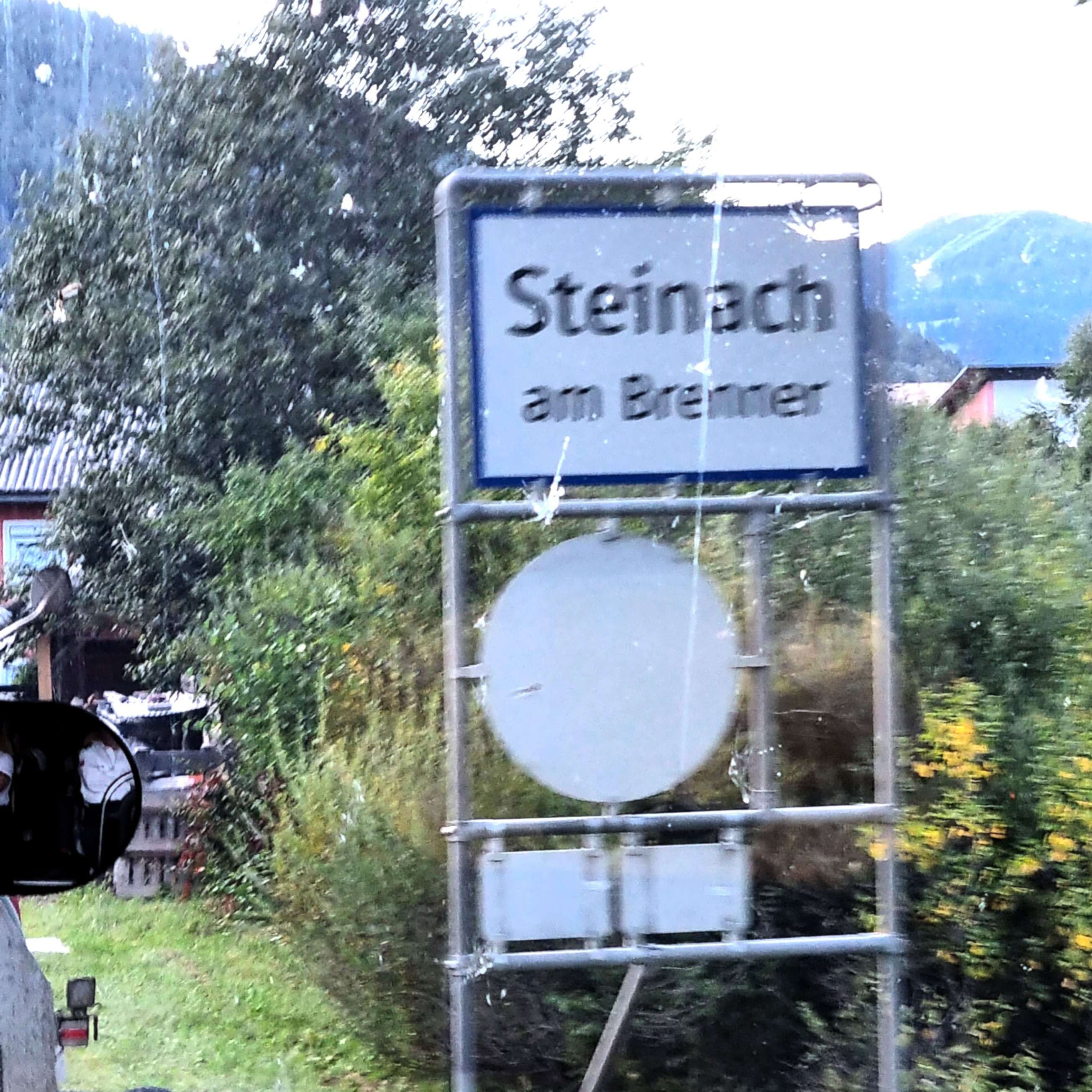Ortsschild mit der Aufschrift 'Steinach am Brenner' an einer Straße, umgeben von Büschen und Bäumen.