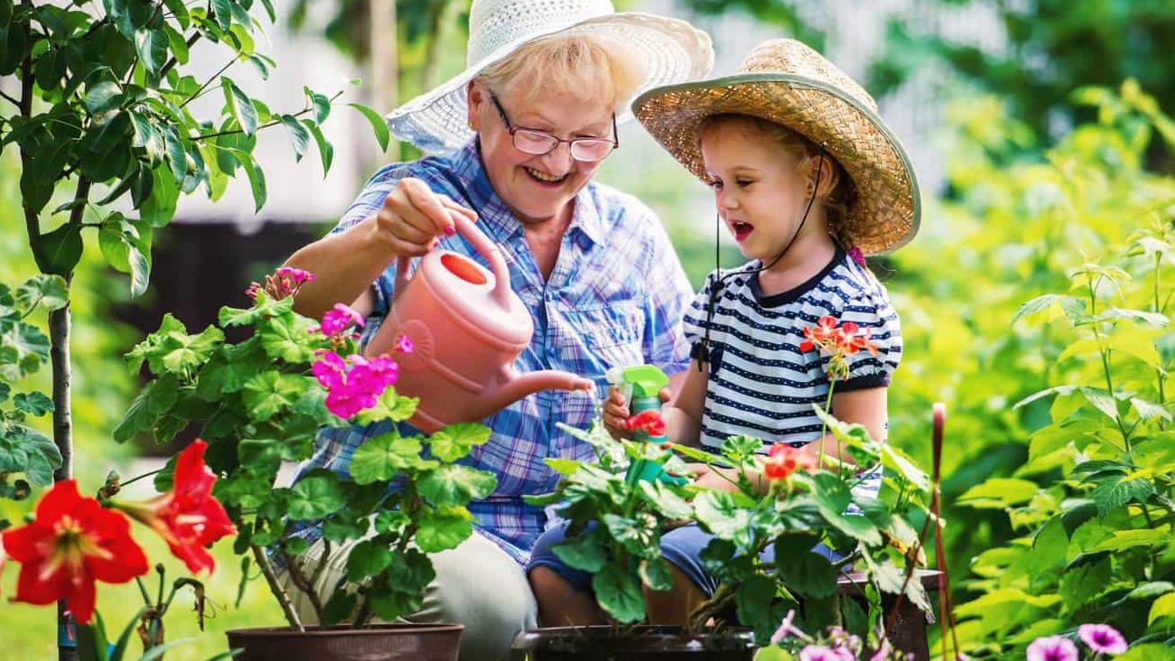 Eine ältere Dame gießt Blumen mit einem jungen Mädchen in einem bunten Garten.