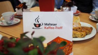 Eine Gruppe verschiedener Personen sitzt an einer Kaffee-Tafel. Eine Helferin der Lohner Malteser reicht Kaffee zum Kuchen.