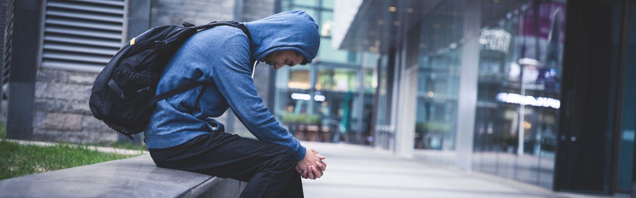 Junger Mann sitzt mit Kapuzenpullover, Kopf gesenkt, auf einer Mauer