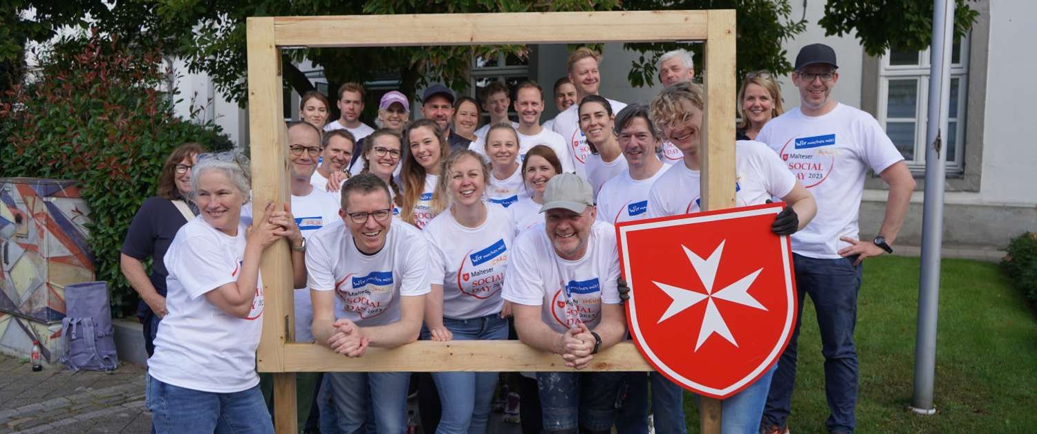 Eine Gruppe von Menschen mit Malteser Social Day T-Shirts halten einen großen Holzrahmen