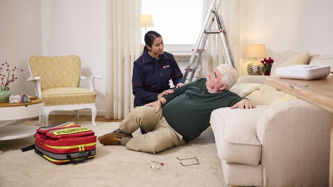 Malteser Mitarbeiterin kniet vor einem im Wohnzimmer gestürzten Senior und bietet ihre Hilfe an.