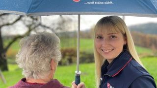 Malteser Mitarbeiterin hält einen Regenschirm über eine Seniorin
