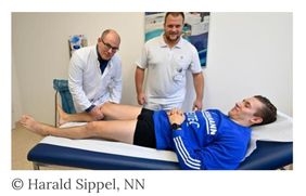 Zwei Mediziner untersuchen das Bein eines Patienten im Trikot, der auf einer Liege liegt.
