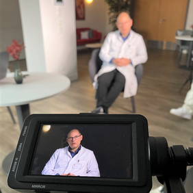 Prof. Dr. Tischer in weißem Kittel bei einem Videodreh, sitzend, im Kameradisplay deutlich im Portrait erkennbar.
