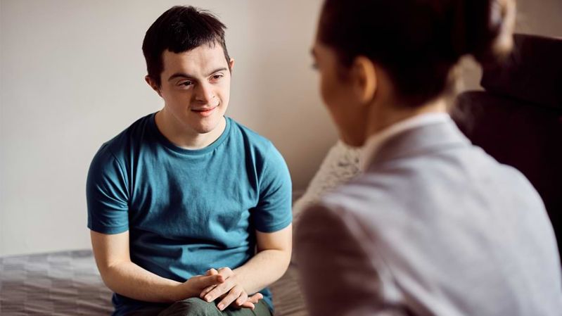 Ein junger Mann mit Trisomie 21 sitzt einer Frau gegenüber und hört ihr zu.