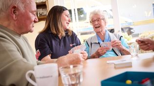 Mehrere Personen sitzen gemeinsam an einem Tisch und spielen Karten. Auf dem Tisch stehen Gläser und eine Tasse. Im Hintergrund fällt durch ein großes Fenster Tageslicht in den Raum, draußen ist ein Fahrzeug der Malteser zu erkennen.