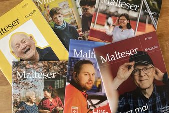 Sechs Ausgaben des „Malteser Magazin“ liegen überlappend auf Holz, mit verschiedenfarbigen Covern und teils unscharfen Personenfotos.