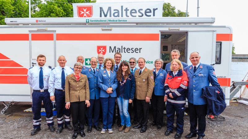 Eine Gruppe von Personen in verschiedenen Uniformen steht vor einem Sanitäts-Anhänger der Malteser.