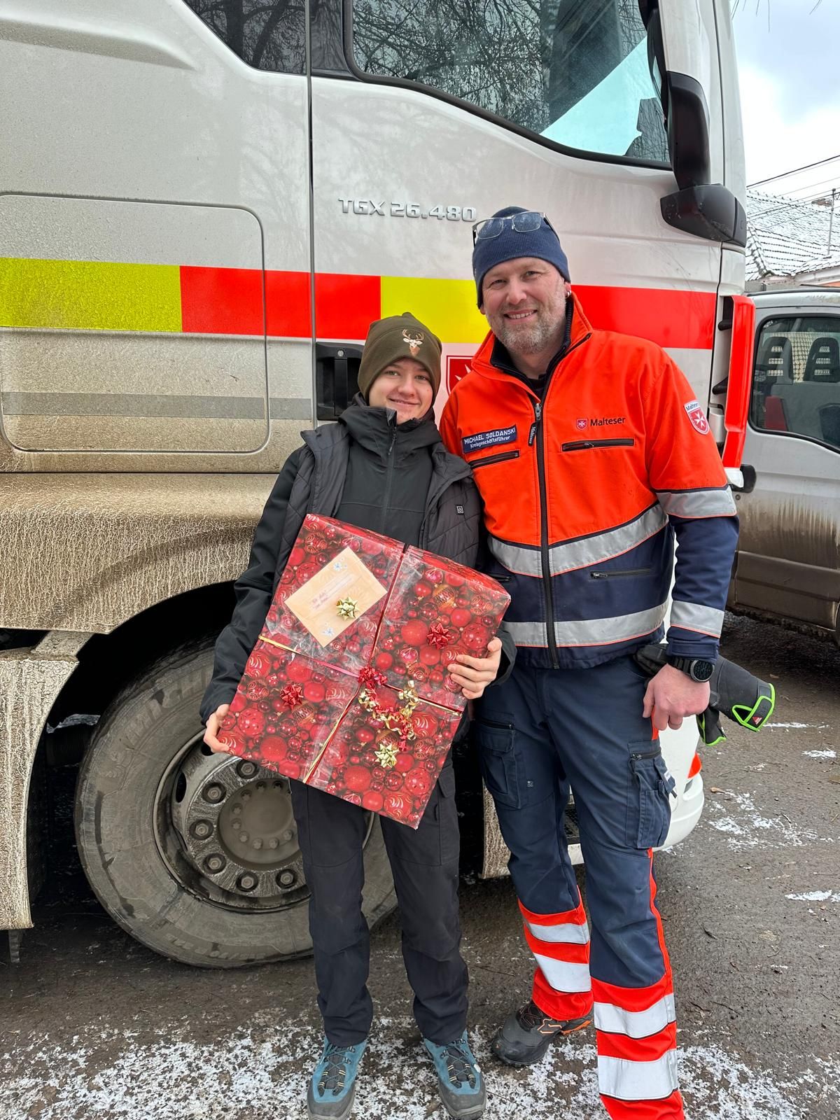 Mann in Malteser Einsatzkleidung und Junge in blauer Jacke mit einem großen Weihnachtspaket stehen neben einem Malteser Lkw