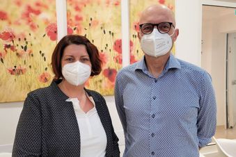 Zwei Personen mit FFP2-Masken stehen in einem Wartebereich vor Blumenbildern.