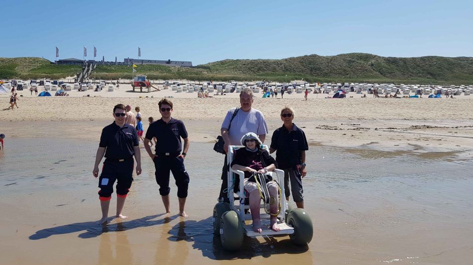 Vier Personen stehen am Ufer eines Strandes, eine Frau sitzt in einem Strandrollstuhl.