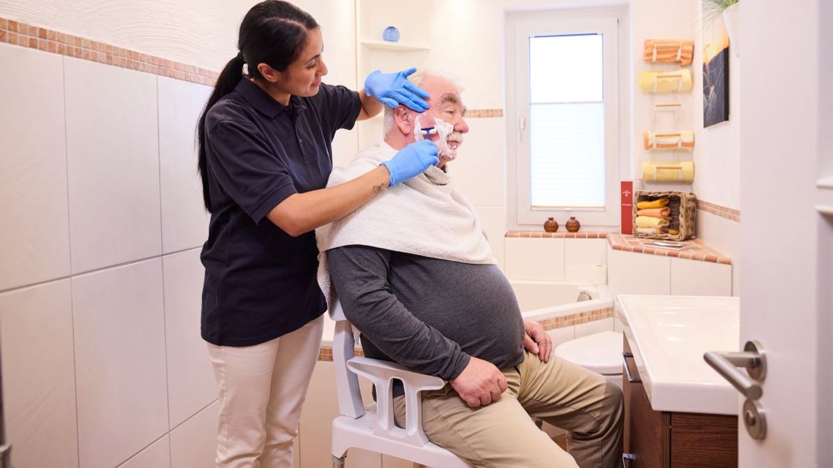 Eine Mitarbeiterin der ambulanten Pflege der Malteser rasiert einen älteren Mann im Badezimmer.