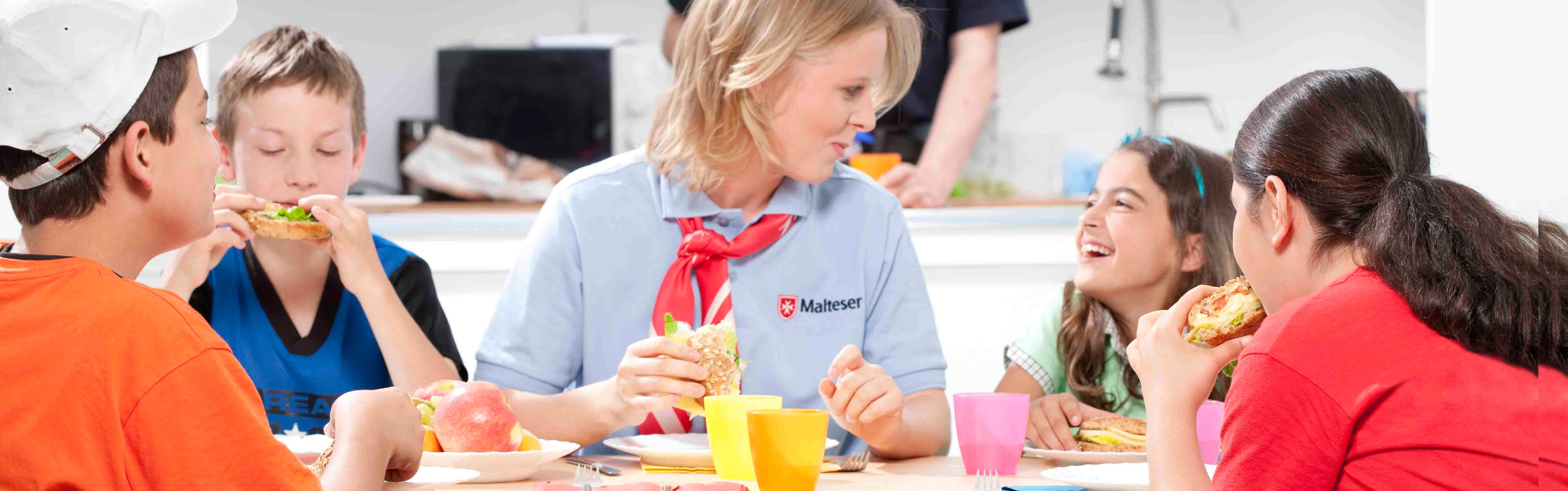 Ehrenamtliche Helferin isst mit Kindern am Tisch.