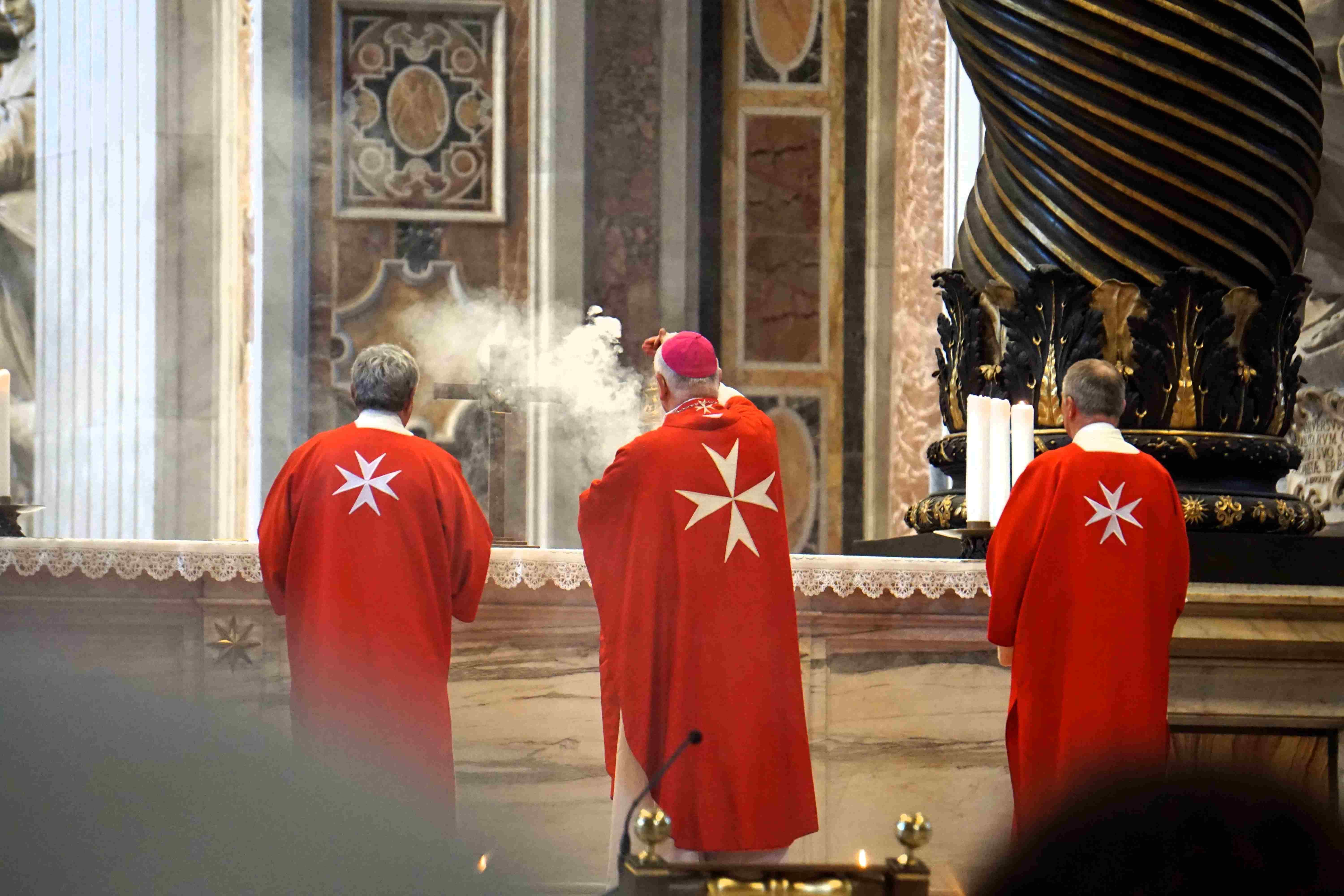 Drei Personen in roten Gewändern mit weißem Malteserkreuz stehen in einer Kirche, eine Person schwenkt Weihrauch.