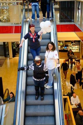 Zwei Malteserinnen begleiten eine Frau im Alterssimulationsanszug eine Rolltreppe herunter