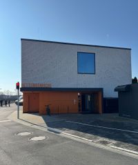 Ein modernes Gebäude mit einer hellen Klinkerfassade und einem großen rechteckigen Fenster im oberen Bereich. Im Erdgeschoss befindet sich ein orangefarbener Eingangsbereich mit der gut sichtbaren Beschriftung „Rettungswache“. Vor dem Gebäude steht ein Fahrrad. Links am Straßenrand ist eine rote Fußgängerampel zu sehen und im Hintergrund stehen einige Personen. Der Himmel ist klar und blau.