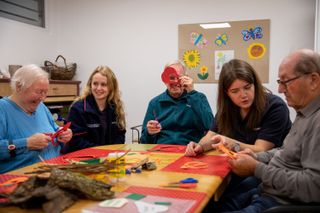 3 ältere Menschen sitzen zusammen mit 2 Malteserinnen an einem Tisch und schneiden zum Training der Feinmotorik etwas aus.