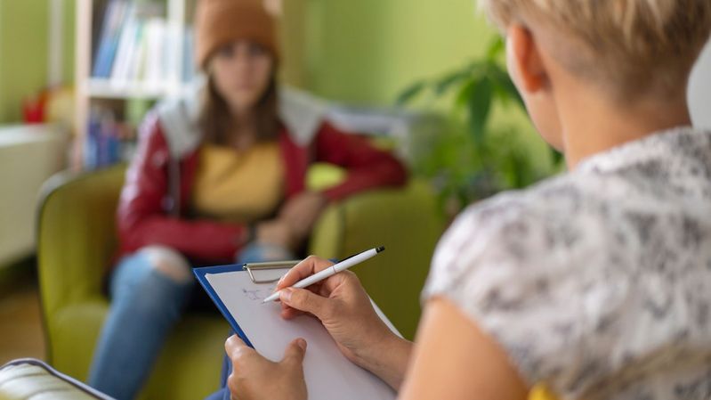 Junge Frau mit bunter Kleidung sitzt einer Psychologin mit Klemmbrett gegenüber. 