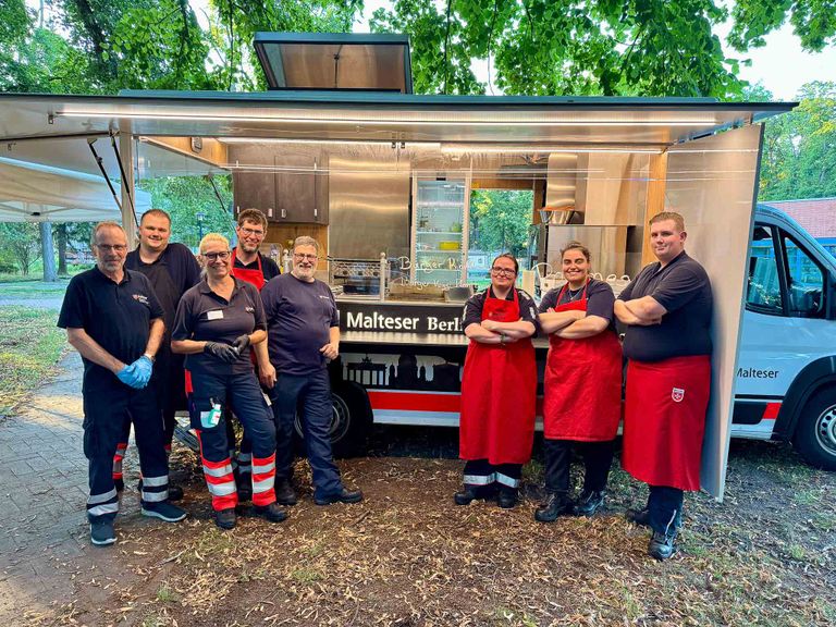 Das Foto zeigt den Gerätewagen Verpflegung mit acht Malteser Helfenden davor.