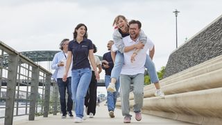 Eine Gruppe junger Menschen, jeglichen Aussehens und Geschlechts, spaziert lachend auf einer ausgebauten Flusspromenade entlang. Vorne wird eine Frau von einem Mann Huckepack getragen.