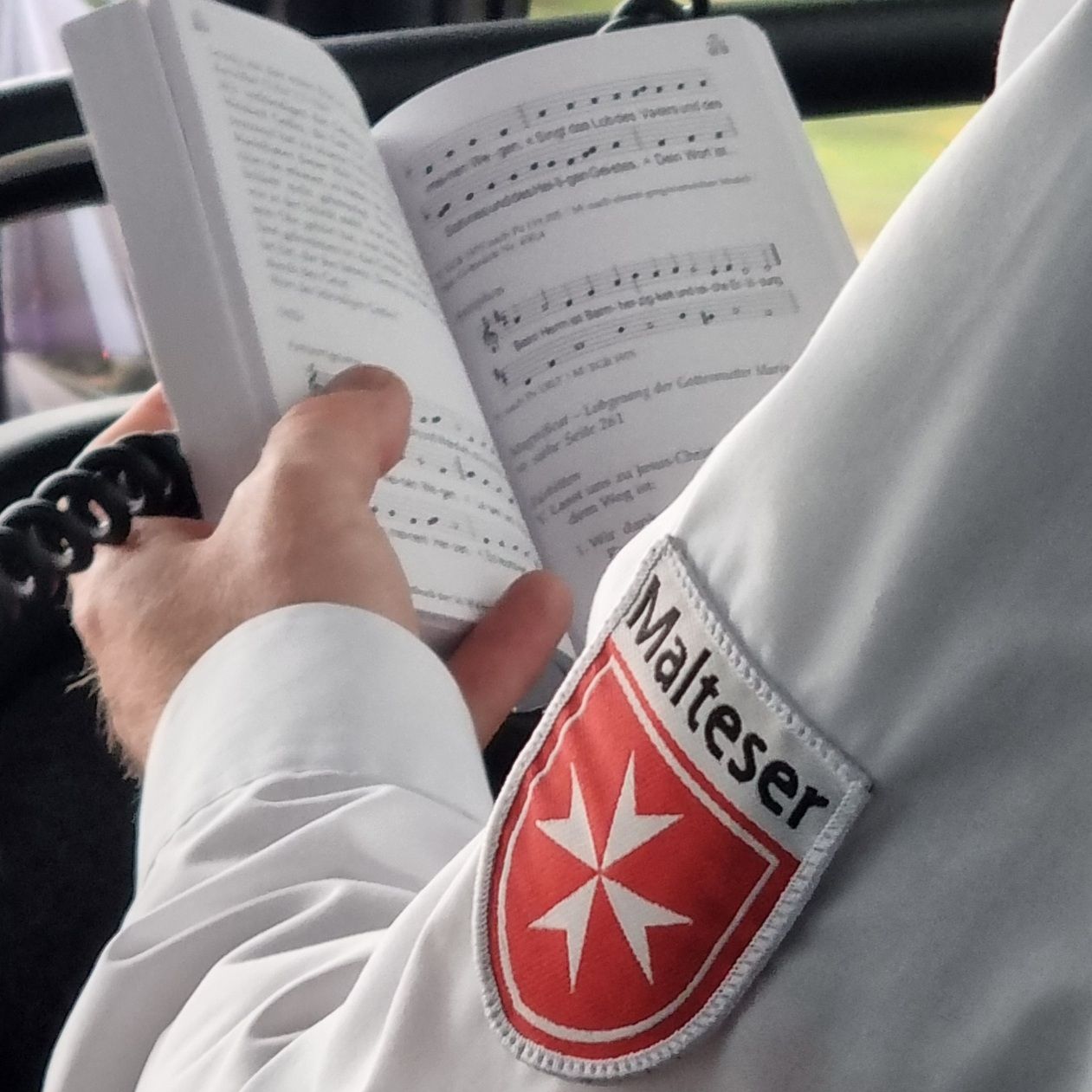 Person in weißem Hemd mit Malteser-Logo auf dem Ärmel hält ein aufgeschlagenes Buch mit Noten und Text.