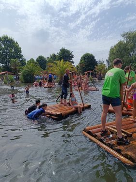 Mehrere Flöße treiben auf einem kleinen See. Kinder und Jugendliche sitzen und stehen drauf oder schieben die Flöße aus dem Wasser heraus an. Im Hintergrund sieht man Palmen und Bäume.