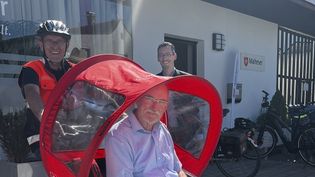 Mann in hellblauem Hemd wird in der roten Malteser Rikscha von einem Mann mit Fahrradhelm und oranger Weste gefahren, daneben steht ein weiterer Mann