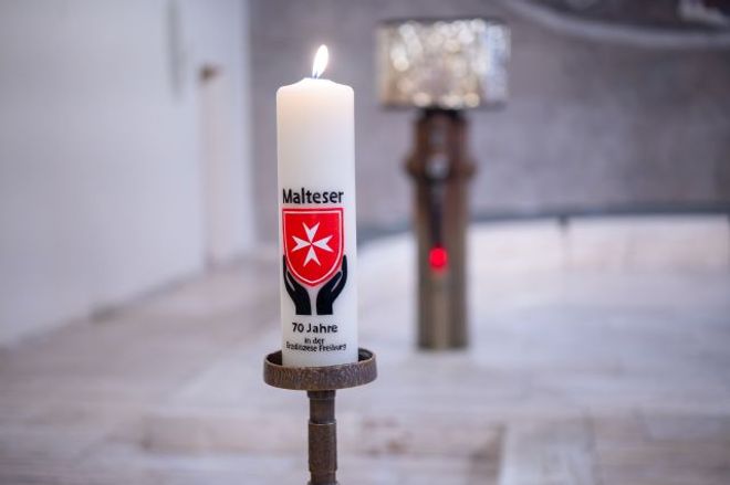 Eine brennende Kerze mit Malteser-Logo steht in einer Kirche vor einem Altar.