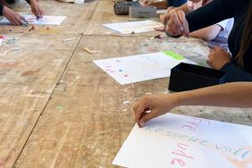 Mehrere Personen gestalten an einem Holztisch bunte Zeichnungen auf Papier. Eine Person malt mit einem Stift auf einem Blatt, während weitere Personen am Tisch arbeiten.